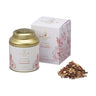 A gold tin container of Ginger & Pomegranate Herbal Tea next to its white box with floral patterns. Loose tea with ginger and pomegranate seeds is also shown.