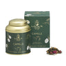 A green La Via del Tè tea box and a gold-colored tin with the name 'Camille' on it, alongside loose leaf green tea and a small pile of dried tea leaves and flowers.