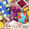 Three colorful tea bags with fruit-themed designs on a wooden surface with fruits around.
