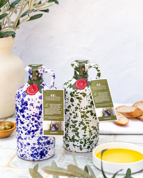 Two decorative ceramic olive oil bottles with hand painted patterns on a table with bread and olives.