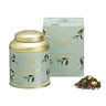 A gold-colored metal tea tin with a floral pattern and the name 'MARGUERITE' on it, alongside a matching box and a small pile of dried tea leaves.