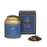 A gold aluminum tin of Segreto dei Medici loose leaf tea with a blue label, accompanied by a pile of dried tea leaves and a box with matching design.