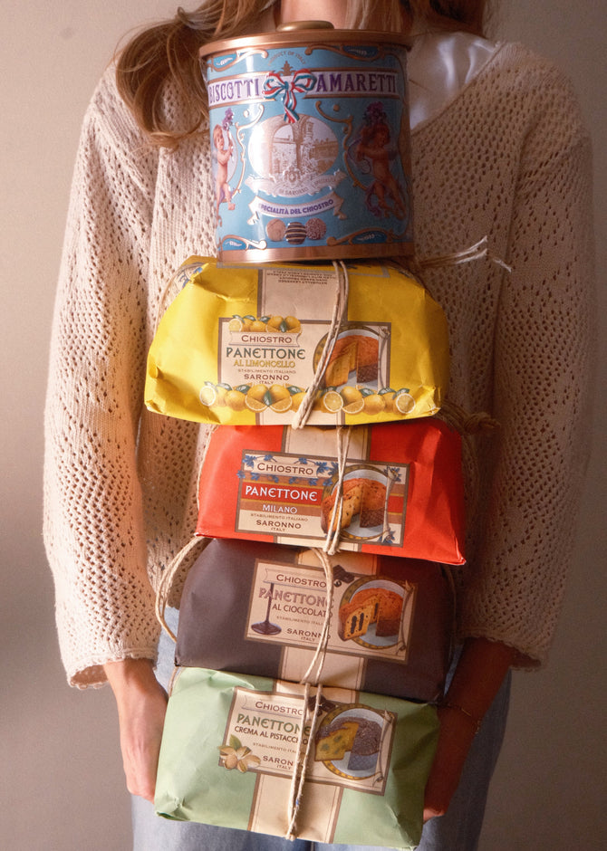 A person wearing a cream knit cardigan holds a stacked bundle of artisanal Panettone boxes wrapped in colorful paper (blue, yellow, orange, brown, and green) tied together with twine.